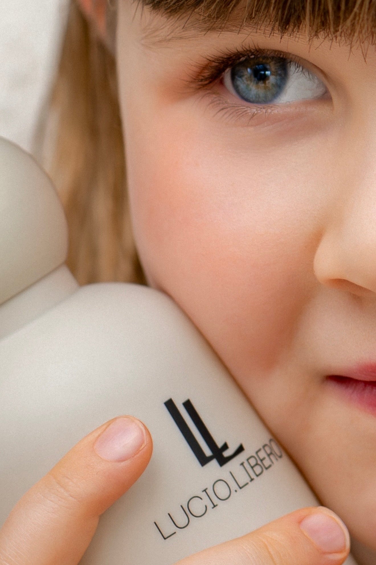 A close-up lifestyle shot of a young girl with brown hair and blue eyes holding a matte off-white bottle of LUCIO LIBERO hair product against her cheek.