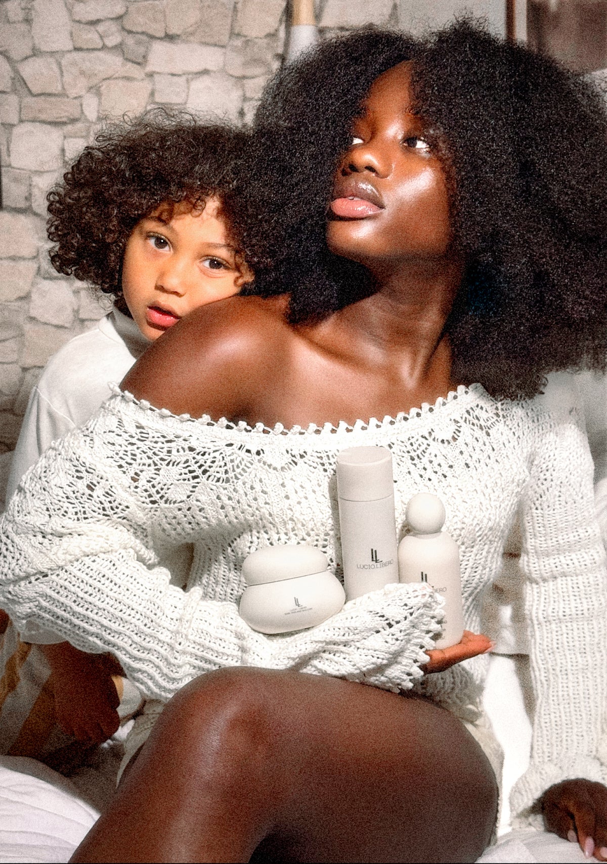Mother with natural curly hair holding her curly-haired child, cradling LUCIO.LIBERO Hair & Skin Balm against a stone wall.